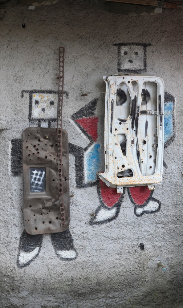 murales su una parete di cemento con due figure antropomorfe con faccine e applicazioni metalliche sul disegno, tra cui uno sportello