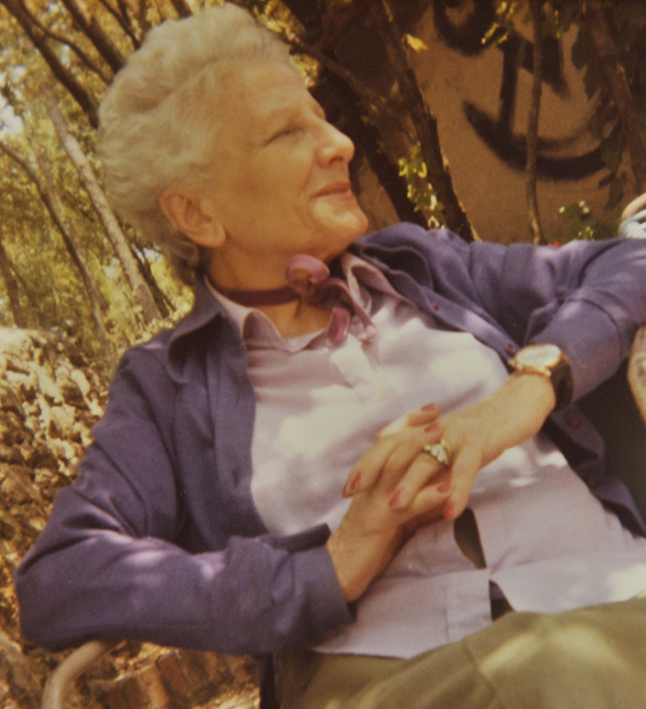Bettina anziana siede in uno spazio all’aperto del Fuseum. Ha le mani giunte, i gomiti appoggiati ai braccioli di una seduta e guarda sorridente alla sua sinistra