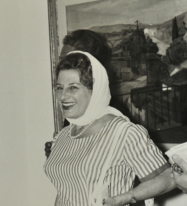 Bettina Fuso sorride verso l’obiettivo mentre osserva un suo dipinto. Indossa un vestito estivo a righe, una collana di perle e ha un foulard in testa