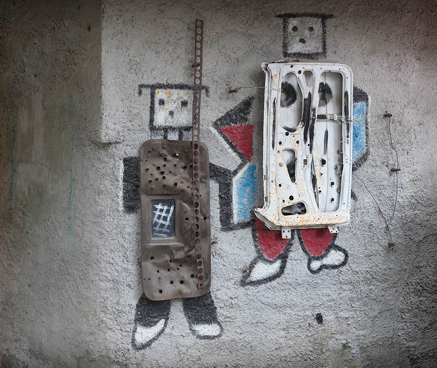 Pittura murale che rappresenta due figure squadrate, che assomigliano a dei robot. Ognuna ha degli inserti meccanici, come lamiere e pezzi di metallo.
