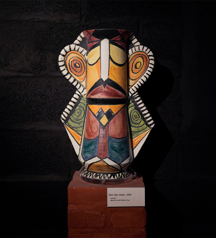 Ceramica smaltata di Brajo Fuso dai colori vivaci posta su un piedistallo. Ripropone una figura umana, con dei baffi dipinti sul viso. I motivi geometrici lungo il busto ricalcano un completo giacca e cravatta.